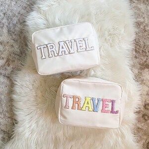 May include: Two off-white travel pouches with the word "TRAVEL" embroidered on the front. One pouch has silver embroidery, and the other has colorful pastel embroidery. The pouches are rectangular and sit on a white fluffy surface.