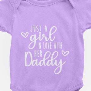 Może przedstawiać: Jasno-fioletowy śpioszek dla niemowlęcia z białym napisem "Just a girl in love with her Daddy" z symbolem serca przed i po tekście.