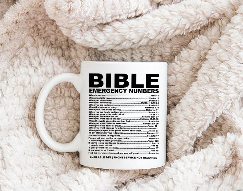 May include: White ceramic mug with a black design that says "BIBLE EMERGENCY NUMBERS" and lists bible verses for different situations. The mug is on a white fuzzy surface.