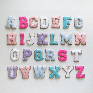 Puede incluir: Un conjunto de letras del alfabeto bordadas en colores pastel, incluyendo rosa, azul y lavanda. Cada letra tiene un borde dorado y una textura esponjosa, dispuestas sobre una superficie blanca. Las letras son de estilo universitario cl&aacute;sico.
