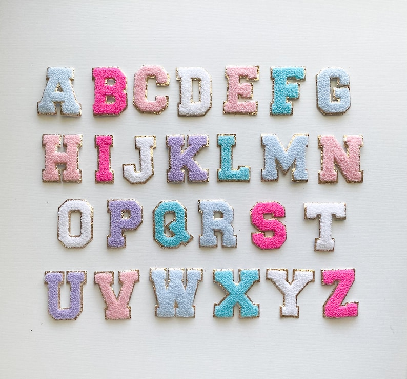 Puede incluir: Un conjunto de letras del alfabeto bordadas en colores pastel, incluyendo rosa, azul y lavanda. Cada letra tiene un borde dorado y una textura esponjosa, dispuestas sobre una superficie blanca. Las letras son de estilo universitario cl&aacute;sico.