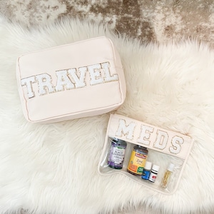 Könnte beinhalten: Zwei rosa Reißverschlusstaschen mit durchsichtigen Vinyl-Einsätzen. Die größere Tasche hat das Wort "TRAVEL" in weißen Buchstaben mit goldenen Glitzerakzenten. Die kleinere Tasche hat das Wort "MEDS" in weißen Buchstaben mit goldenen Glitzerakzenten. Die kleinere Tasche enthält mehrere Flaschen mit Pillen und ätherischen Ölen.