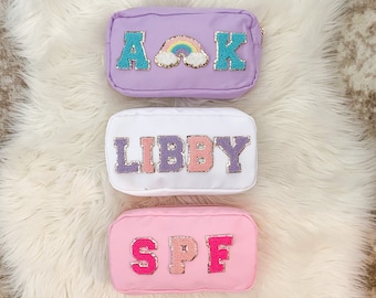 Könnte beinhalten: Drei kleine Reißverschlusstaschen mit verschiedenen Stickereien. Die obere Tasche ist lila mit den Buchstaben "AAK" und einem Regenbogen. Die mittlere Tasche ist weiß mit dem Namen "LIBBY" in rosa und lila Buchstaben. Die untere Tasche ist rosa mit den Buchstaben "SPF" in rosa und weißen Buchstaben.