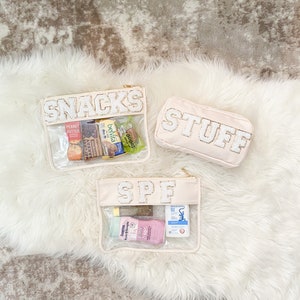 Könnte beinhalten: Drei transparente Reißverschlusstaschen mit weißen Stoffaußenflächen. Die Taschen sind mit "SNACKS", "STUFF" und "SPF" beschriftet. Die Taschen enthalten verschiedene Snacks, Toilettenartikel und Sonnencreme.