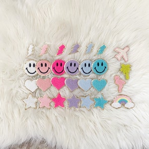 Puede incluir: Colecci&oacute;n de parches coloridos y bordados en varias formas, incluyendo caras sonrientes, corazones, estrellas, rayos y un arco&iacute;ris. Cada parche tiene un borde dorado sobre un fondo blanco y esponjoso.
