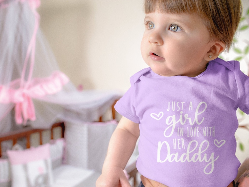 Może przedstawiać: Dziecko w liliowym body z napisem "Just a girl in love with her Daddy" w białym kolorze.