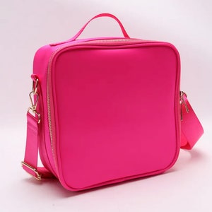 Puede incluir: Una bolsa de almuerzo rosa brillante con un asa superior y una correa para el hombro desmontable. La bolsa tiene forma cuadrada con una cremallera dorada y herrajes dorados. La bolsa est&aacute; hecha de un material liso de color s&oacute;lido.