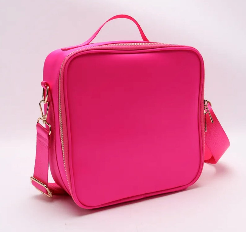 Puede incluir: Una bolsa de almuerzo rosa brillante con un asa superior y una correa para el hombro desmontable. La bolsa tiene forma cuadrada con una cremallera dorada y herrajes dorados. La bolsa est&aacute; hecha de un material liso de color s&oacute;lido.