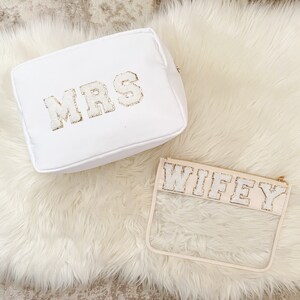 May include: Two white cosmetic bags with gold glitter lettering. One bag is a white fabric pouch with the word "MRS" on it. The other bag is a clear plastic pouch with the word "WIFEY" on it.