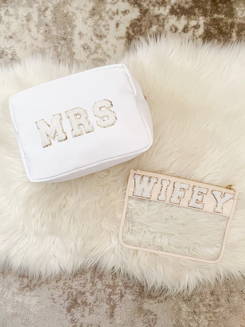 May include: Two white cosmetic bags with gold glitter lettering. One bag is a white fabric pouch with the word "MRS" on it. The other bag is a clear plastic pouch with the word "WIFEY" on it.