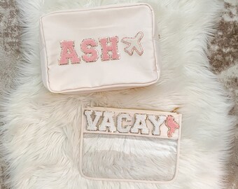 Könnte beinhalten: Zwei Kosmetiktaschen für unterwegs. Eine ist eine rechteckige, hellrosa Tasche mit dem Namen "ASH" in rosa und goldenen Buchstaben und einer Flugzeuggrafik. Die andere ist eine transparente Tasche mit dem Wort "VACAY" in weißen und goldenen Buchstaben und einer rosa Grafik.