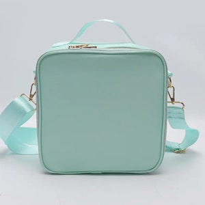 Puede incluir: Bolso de mano cuadrado verde menta con asa superior y bandolera extra&iacute;ble. El bolso presenta herrajes dorados, incluyendo cremalleras y cierres. La correa y el asa est&aacute;n hechos de un material verde menta a juego.