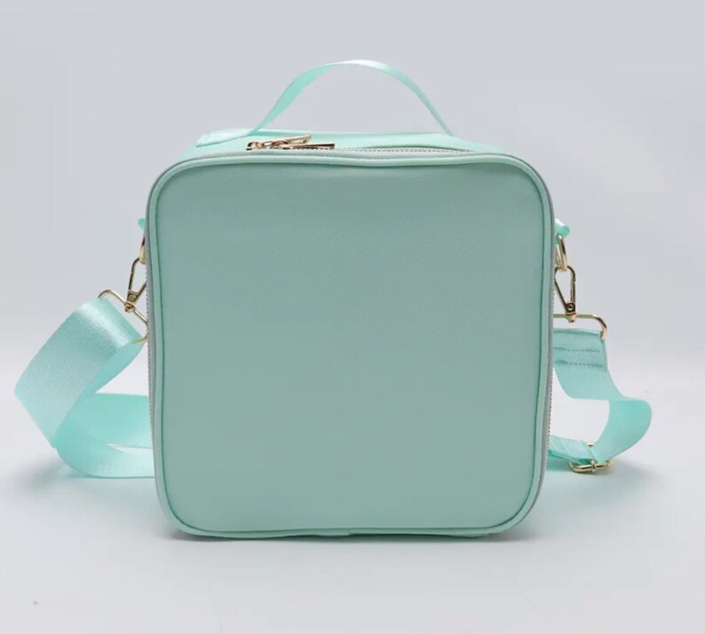 Puede incluir: Bolso de mano cuadrado verde menta con asa superior y bandolera extra&iacute;ble. El bolso presenta herrajes dorados, incluyendo cremalleras y cierres. La correa y el asa est&aacute;n hechos de un material verde menta a juego.