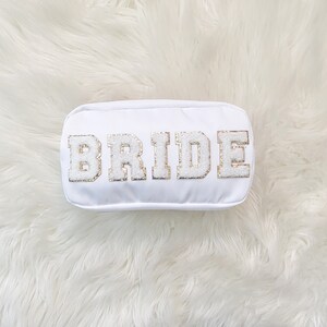 May include: White makeup bag with the word "BRIDE" in gold glitter letters.