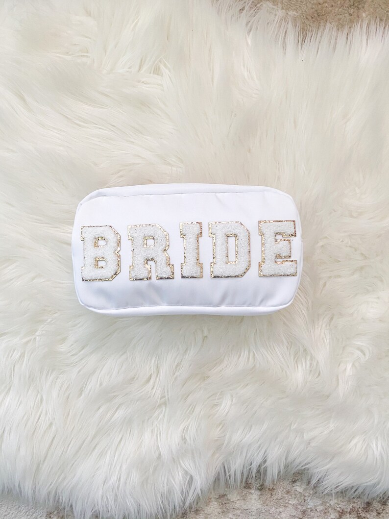 May include: White makeup bag with the word "BRIDE" in gold glitter letters.