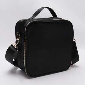 Puede incluir: Un bolso negro de forma cuadrada con un asa superior y una correa para el hombro desmontable. El bolso tiene una cremallera y herrajes dorados. El bolso est&aacute; hecho de un material liso y mate.