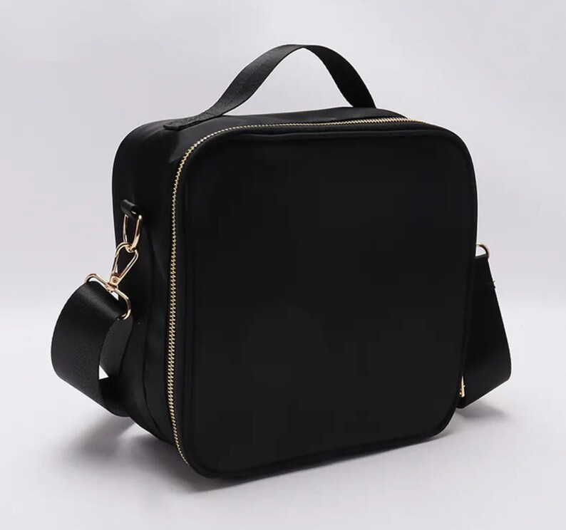 Puede incluir: Un bolso negro de forma cuadrada con un asa superior y una correa para el hombro desmontable. El bolso tiene una cremallera y herrajes dorados. El bolso est&aacute; hecho de un material liso y mate.