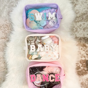 Puede incluir: Tres bolsas transparentes con cremallera de plástico con exteriores de tela de diferentes colores. Las bolsas están etiquetadas con "SWIM", "BABY" y "DANCE" con letras de colores. Las bolsas están llenas de varios artículos.
