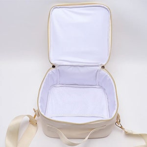Puede incluir: Bolsa de almuerzo crema abierta, con cremallera dorada e interior blanco. La bolsa tiene un bolsillo de malla blanco en el interior y una correa para el hombro color crema. La bolsa es de forma cuadrada con esquinas redondeadas.