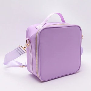 Puede incluir: Una bolsa de almuerzo de color lavanda con un asa superior y una correa para el hombro desmontable. La bolsa tiene forma cuadrada con cremallera y herrajes dorados. La correa y el asa son del mismo color lavanda que la bolsa.