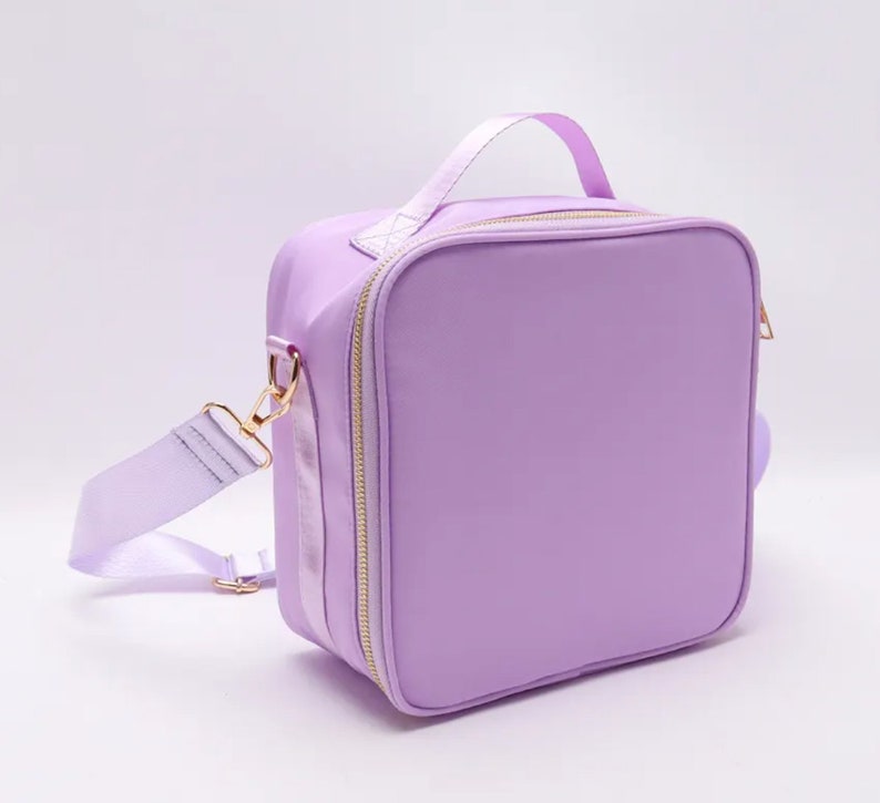 Puede incluir: Una bolsa de almuerzo de color lavanda con un asa superior y una correa para el hombro desmontable. La bolsa tiene forma cuadrada con cremallera y herrajes dorados. La correa y el asa son del mismo color lavanda que la bolsa.