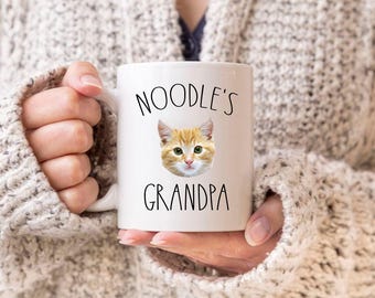 May include: A white ceramic mug featuring a graphic of an orange tabby cat face centered between the black text NOODLE'S and GRANDPA. The mug has a smooth finish and a curved handle on the left side, designed for holding a warm beverage comfortably.