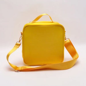 Puede incluir: Una bolsa de almuerzo amarilla brillante, cuadrada, con una correa para el hombro a juego y un asa superior. La bolsa tiene herrajes met&aacute;licos dorados y un cierre de cremallera. La correa es ajustable y extra&iacute;ble.