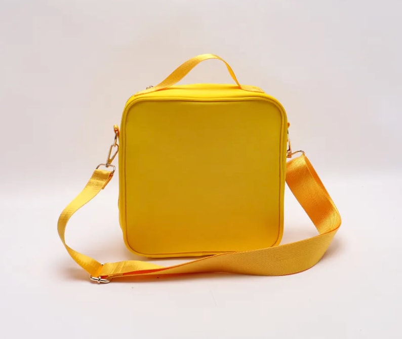 Puede incluir: Una bolsa de almuerzo amarilla brillante, cuadrada, con una correa para el hombro a juego y un asa superior. La bolsa tiene herrajes met&aacute;licos dorados y un cierre de cremallera. La correa es ajustable y extra&iacute;ble.