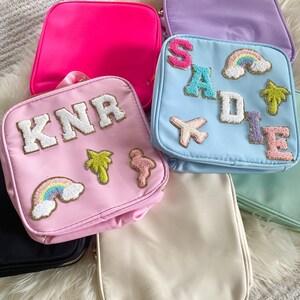 Puede incluir: Una colecci&oacute;n de neceseres coloridos en varios tonos, incluyendo rosa, morado y azul. Cada neceser presenta parches bordados con letras, arco&iacute;ris y otros dise&ntilde;os. Los neceseres est&aacute;n dispuestos sobre una superficie blanca y esponjosa.