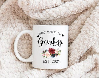 Puede incluir: Una taza de cerámica blanca con el texto en negro PROMOTED TO Grandma EST. 2021, dos pequeños corazones negros y un colorido arreglo floral de flores de color rojo oscuro, rosa y azul con hojas verdes impreso en la parte frontal.