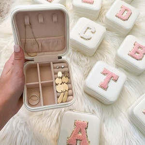 May include: White square jewelry boxes with a pink initial letter on the lid. One box is open, revealing a necklace, earrings, and rings. The boxes are on a white fluffy surface.