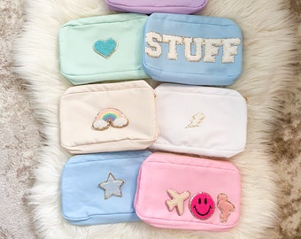 Könnte beinhalten: Ein Set aus sieben bunten Reißverschlusstaschen mit verschiedenen gestickten Aufnähern. Die Taschen sind in den Farben Lavendel, Hellblau, Hellgrün, Rosa, Weiß und Beige. Die Aufnäher beinhalten ein Smiley, ein Herz, das Wort "STUFF", einen Regenbogen, einen Blitz, einen Stern und ein Flugzeug mit einem Smiley.