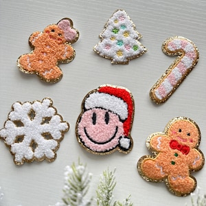 May include: Six felt Christmas-themed patches with gold glitter trim. The patches include a gingerbread person, a Christmas tree, a candy cane, a snowflake, a smiling face with a Santa hat, and another gingerbread person.