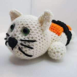 May include: White crocheted cat plush toy with black eyes and whiskers. The cat is wearing a crocheted orange and black sushi roll costume.