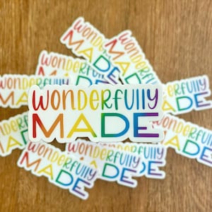 Puede incluir: Una colección de pegatinas blancas con las palabras "Wonderfully Made" en una fuente de gradiente arcoíris. Las pegatinas están esparcidas sobre una superficie de madera, con el texto en varios tamaños y orientaciones.