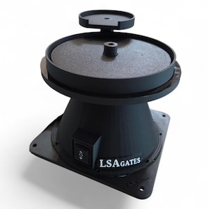 Puede incluir: Dispositivo negro impreso en 3D con una parte superior circular y una base cuadrada. El dispositivo tiene una superficie texturizada en la parte superior y una pequeña plataforma encima. El texto "LSA GATES" está impreso en el lateral.