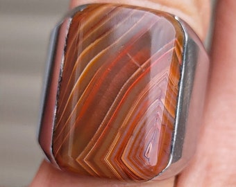 May include: A silver ring with a rectangular, brown agate stone. The stone has a swirling pattern of white, orange, and red.