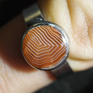 May include: A silver ring with a round, orange and white agate stone set in the center. The stone has a swirling, layered pattern.