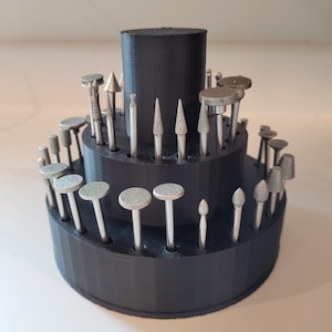 May include: A tiered black organizer holds various silver and gray rotary tool bits. The bits have different shapes, including cones, cylinders, and rounded tips. The organizer is made of black plastic and sits on a white surface.