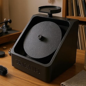 May include: A black record cleaning machine with a circular cleaning pad and a brush sits on a wooden table. A turntable and records are in the background. The machine has the text "ISAGATES" on the front.