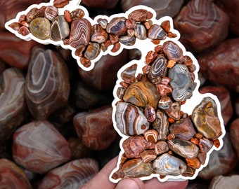 May include: A hand holds a sticker in the shape of the state of Michigan, filled with colourful, polished stones. The stones are various sizes and shapes, with brown, red, and white patterns. The background is a blurred image of similar stones.