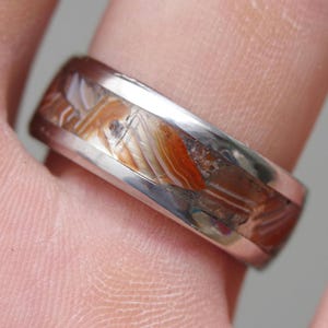 May include: A silver ring with a brown and white agate inlay. The ring is worn on a person's finger.