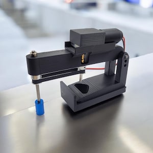 Puede incluir: Una máquina negra impresa en 3D con un componente cilíndrico azul. El dispositivo tiene una base rectangular, un mecanismo deslizante y un motor. Se ve un cable rojo. La máquina está sobre una superficie metálica.