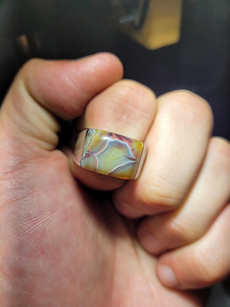 May include: A silver ring with a square-shaped agate gemstone. The agate has a swirling pattern of yellow, brown, red, and white.