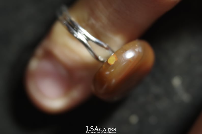 May include: A close-up of a ring with a brown and orange gemstone. The ring is made of silver and has a simple band.