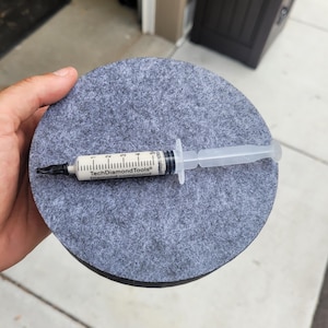 Puede incluir: Una jeringa transparente llena de una sustancia blanca, colocada sobre un posavasos de fieltro gris. La jeringa tiene marcas en blanco y negro y el texto "TechDiamondTools Diamond compound". El posavasos es redondo.