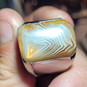 May include: A silver ring with a large, rectangular, agate stone. The stone has a white, brown, and yellow banded pattern.