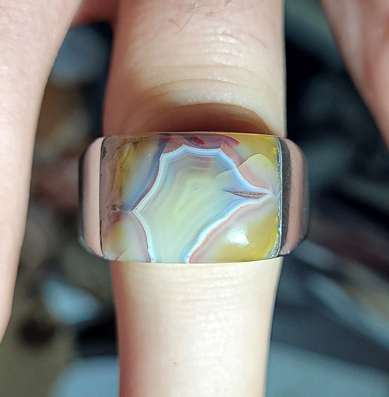 May include: A silver ring with a large, rectangular, multi-colored agate stone set in the center. The stone has a swirling pattern of brown, white, yellow, and blue.