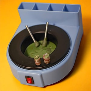 Puede incluir: Un recipiente de cera azul claro con un anillo interior negro, que contiene cera verde y dos herramientas de metal. Dos objetos pequeños y cilíndricos están delante. El recipiente tiene un interruptor y una sección con tres compartimentos.