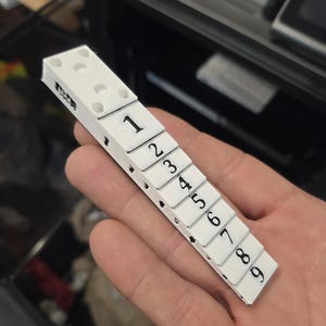 Puede incluir: Una pieza de juego tipo dominó blanca con números negros del 1 al 9. La pieza del juego se sostiene en una mano y los números son claramente visibles. La pieza del juego está hecha de un material blanco con números negros.